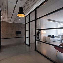 Couloir de bureau moderne avec murs en verre à montants en métaux noirs, sol en béton lisse, plafond à panneaux, et vue sur un espace salon avec canapé clair et lampe sur pied.